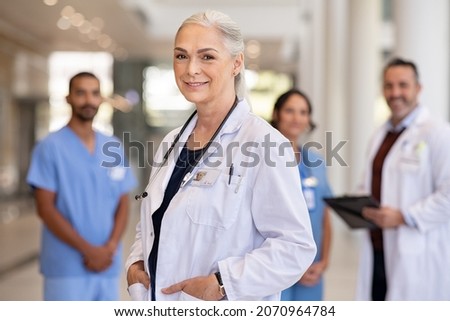 Portrait of mature female doctor on hospital corridor. Confident general practitioner standing in hospital hallway with her healthcare team in background. Successful head physician in private clinic. Royalty-Free Stock Photo #2070964784