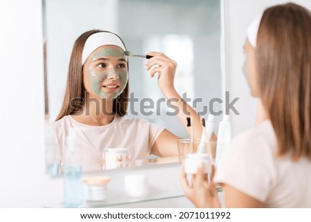 beauty, skin care and people concept - teenage girl with brush applying clay mask to face and looking in mirror at bathroom