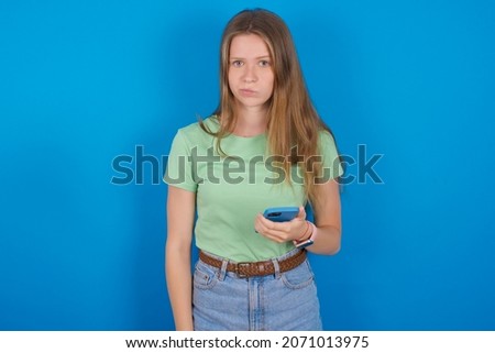 Upset dissatisfied Young caucasian girl wearing green T-shirt over blue background uses mobile software application and surfs information in internet, holds modern mobile hand
