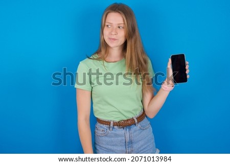 Young caucasian girl wearing green T-shirt over blue background holds new mobile phone and looks mysterious aside shows blank display of modern cellular