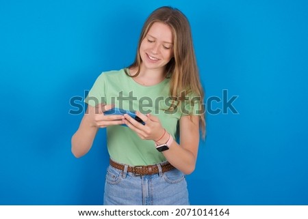 Young caucasian girl wearing green T-shirt over blue background holding in hands cell playing video games or chatting