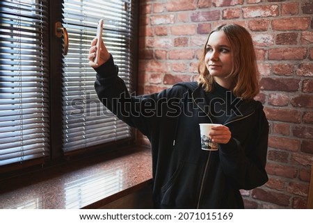 Young woman having video call, talking remotely, taking selfie photo holding smartphone. Girl drinking coffee relaxing while taking break in office standing at window
