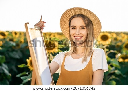 Portrait of a woman who is smiling. She is standing near an easel among sunflowers. She is looking at the camera.