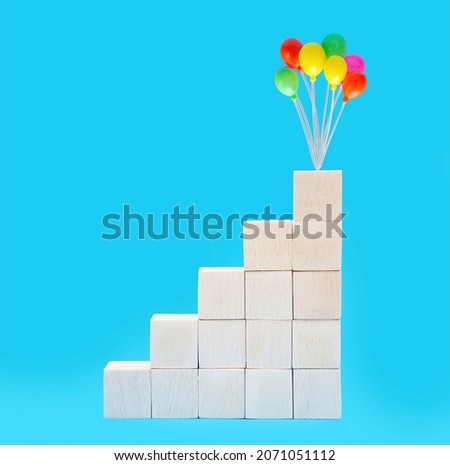 Wooden toy blocks stacked into stairs with colorful party balloons on top isolated on blue background. The concept of taking stairs to success.