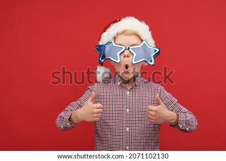 Portrait of happy boy wearing Santa Claus hat and carnaval sunglasses shows thumbs up. Funny child waiting for the holiday on a red background
