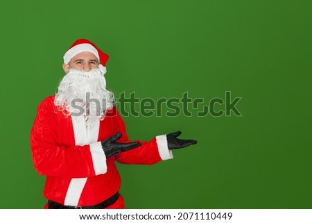 A Caucasian man dressed as Santa Claus is pointing with his open hands to the side where there is an empty space. The background is green.