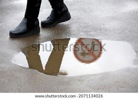 Rain in city, woman in boots walks around the puddle on city street. Road sign forbidding a turn are reflected in water, concept of choosing the wrong way