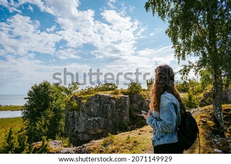 Woman with brown curly hair hiking and taking pictures with phone on the seaside by the Uugu cliff on the Muhu Island near Saaremaa in Estonia during a sunny day