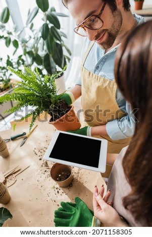 Young florist holding digital tablet with blank screen near smiling colleague with plant in flower shop