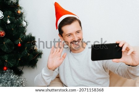 Young handsome positive man in santa hat making video call on mobile phone, congratulating Merry Christmas and Happy New Year sitting celebrating alone at home near decorated Christmas tree.