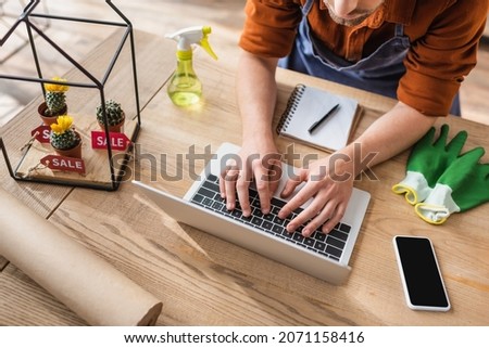 Cropped view of florist using laptop near gloves and cacti with price tags in flower shop