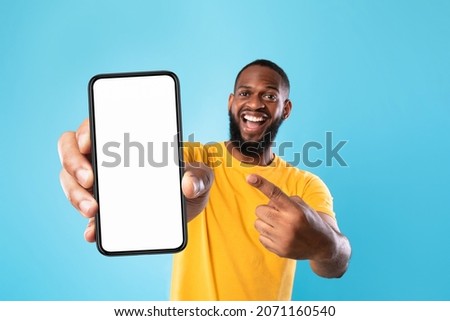 Mobile advertisement. Overjoyed black man pointing at cellphone with empty white screen on blue studio background, mockup for app or website. Cellphone display template Royalty-Free Stock Photo #2071160540