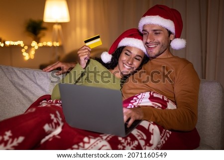 Christmas Shopping. Happy Couple Using Laptop And Credit Card Wearing Santa Hats And Buying Presents Online At Home At Night. Customers Purchasing Gifts Via Computer On Xmas Eve