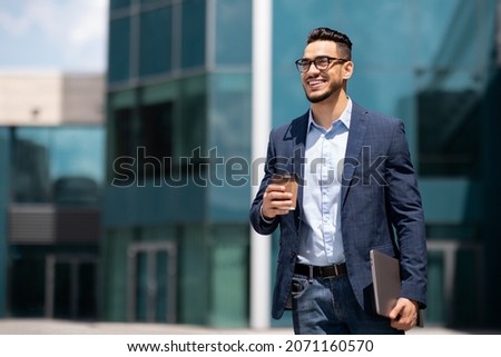 Cheerful middle eastern young businessman in glasses with laptop and coffee to go walking by street towards modern office building, going to job in the morning, panorama with copy space Royalty-Free Stock Photo #2071160570
