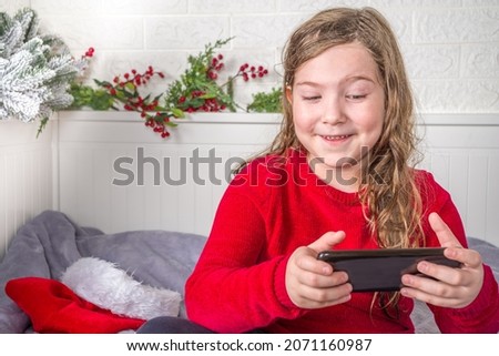 Close up photo of positive kid girl with santa claus hat and red sweater use smartphone, game or chating, christmas congratulation search discounts in house with x-mas decorations
