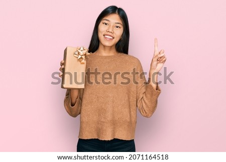 Beautiful young asian woman holding gift surprised with an idea or question pointing finger with happy face, number one 