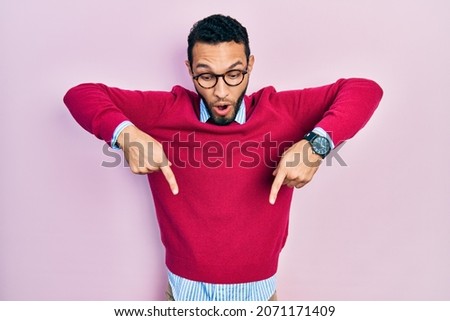 Hispanic man with beard wearing business shirt and glasses pointing down with fingers showing advertisement, surprised face and open mouth 