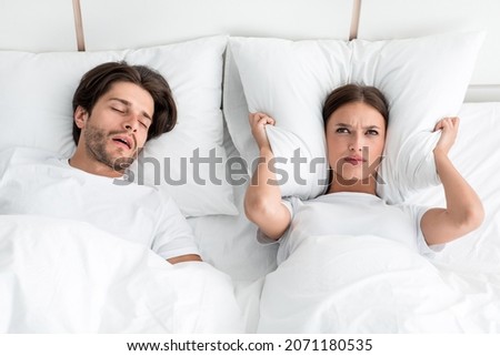 Attractive millennial european guy with stubble sleeps on bed, angry upset wife covers ears with pillow, suffers noise in bedroom interior. Snoring, health and relationship problems, apnea, insomnia Royalty-Free Stock Photo #2071180535