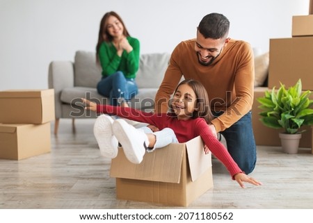 Fooling While Moving. Portrait of happy Arab family having fun together in new apartment, unpacking, smiling dad riding excited little girl in cardboard box in living room, woman sitting on the couch Royalty-Free Stock Photo #2071180562