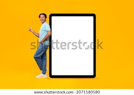 I Recommend. Full body length of cheerful young man leaning on large big white blank digital tablet screen, showing thumbs up sign gesture. Gadget with empty free space for mock up, great mobile offer
