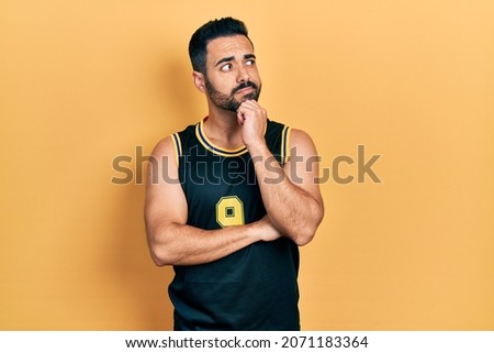 Handsome hispanic man with beard wearing basketball t shirt thinking concentrated about doubt with finger on chin and looking up wondering 