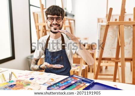 Hispanic man with beard at art studio gesturing with hands showing big and large size sign, measure symbol. smiling looking at the camera. measuring concept. 