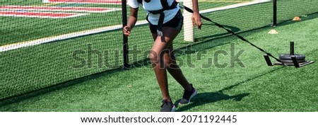 A high school girl track runner is pulling a sled with weights on a turf field next to a protective net during practice.