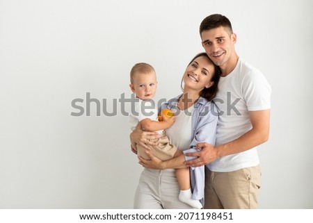 Happy Young Parents Holding Their Cute Toddler Son And Smiling At Camera, Portrait Of Beautiful Cheerful Family Of Three With Little Baby Posing Together Over White Background, Copy Space