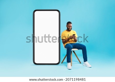Cheerful young black man sitting on chair near big smartphone with empty screen, using mobile device, presenting space for website, advertisement or mobile app, blue studio background Royalty-Free Stock Photo #2071198481