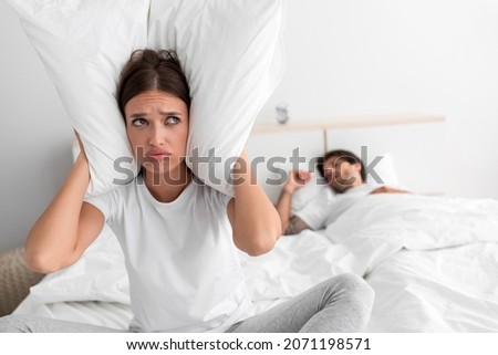 Despaired millennial european wife sits on bed covers ears with pillow and suffers from snoring of sleeping husband in bedroom interior. Intolerable snoring and noise at night, insomnia and apnea Royalty-Free Stock Photo #2071198571