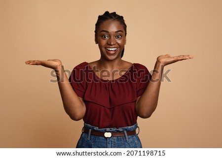 Choice, choosing, balance concept. Confused african american young woman holding something invisible on both palms, trying to choose something, holding hands up as scales, beige studio background Royalty-Free Stock Photo #2071198715