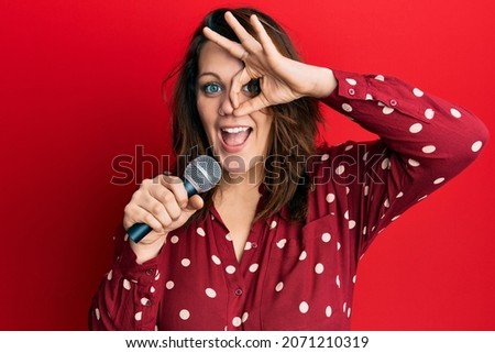 Young caucasian woman singing song using microphone smiling happy doing ok sign with hand on eye looking through fingers 