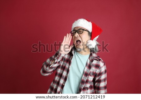 The senior Asian man wears red plaid shirt with Santa Claus hat standing on the red background.