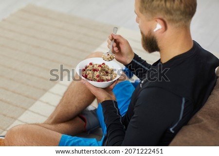 Athlete enjoying healthy meal rich in fiber, protein and vitamins. Fit young man sitting on floor in living room, relaxing after fitness workout, eating natural vegetarian granola, listening to music Royalty-Free Stock Photo #2071222451