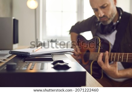 Musician and songwriter writing and playing music. Serious focused man picking guitar strings and thinking of new song while sitting with pencil and notebook by electronic keyboard in home studio