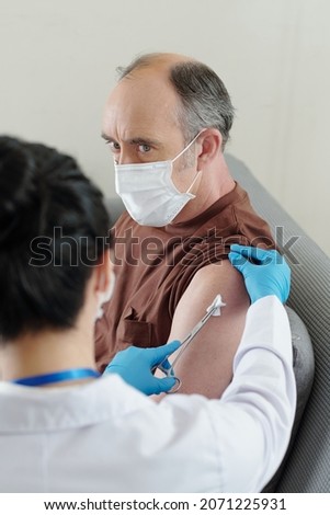 A bit scared senior man looking at doctor cleaning vaccine site on his arm with antiseptic