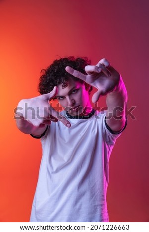 Young man in white T-shirt with picture frame gesture isolated over gradient pink orange background in neon lights. Concept of feelings, youth, fashion, facial expression, emotions, lifestyle, ad
