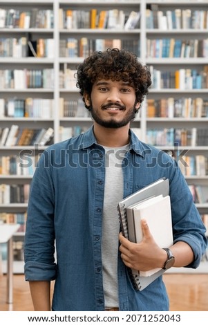 Portrait of satisfied confident smiling young Indian male freelancer or student holding books looking at camera standing in university library. Research for class assignment and exams preparation