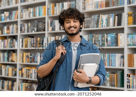 Portrait of cheerful male international Indian student with backpack, learning accessories standing near bookshelves at university library or book store during break between lessons. Education concept Royalty-Free Stock Photo #2071252046
