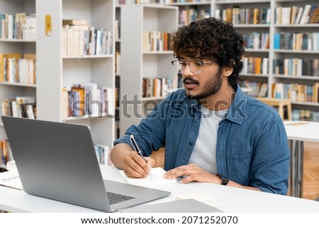 Young Indian male student watching education webinar using laptop writing notes doing research on internet sitting at high school library. Digital technology, communication and studying Royalty-Free Stock Photo #2071252070