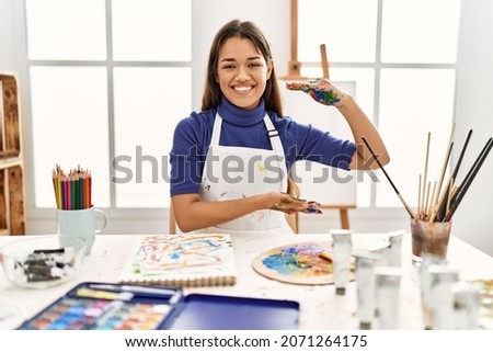 Young brunette woman at art studio with painted hands gesturing with hands showing big and large size sign, measure symbol. smiling looking at the camera. measuring concept. 
