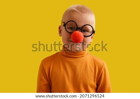 funny boy with a clown nose. Child clown smile shows, copy space area on yellow background. Funny little clown with red nose. Concept birthday, day 1 april, party. Beautiful portrait kid jeste Royalty-Free Stock Photo #2071296524
