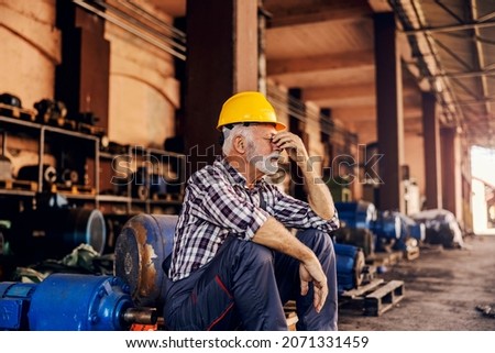 A worried senior factory worker is sitting next to the machines and holding his head. He is hoping the work he has done is fine. A nervous worker at the factory. Royalty-Free Stock Photo #2071331459