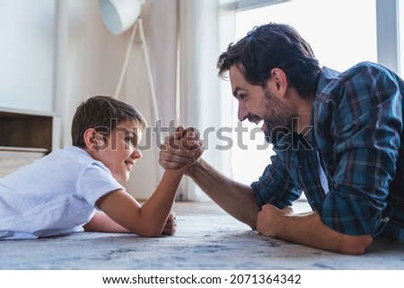 Portrait of a little boy and his dad lying on the floor competing with arm wrestling at home in the living room.