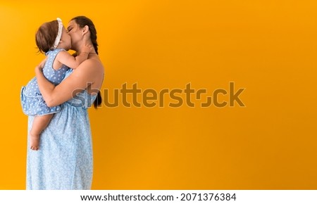 Motherhood, hot summer - Banner young pregnant happy smiling mother woman in blue dress holding little baby daughter toddler sibling in arms looking into eyes kisses on yellow background coppy space