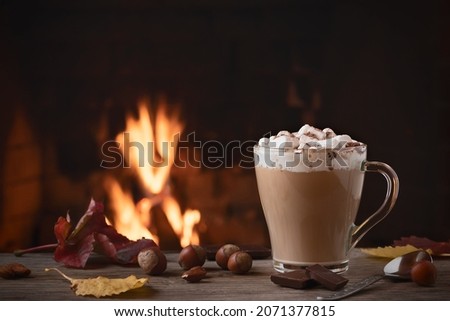 Cocoa with marshmallows and chocolate in a glass mug on a wooden table near a burning fireplace