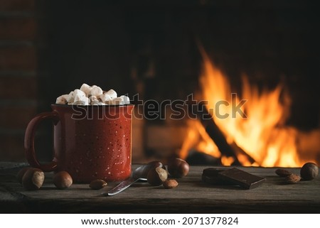 Cocoa with marshmallows and chocolate in a red mug on a wooden table near a burning fireplace Royalty-Free Stock Photo #2071377824