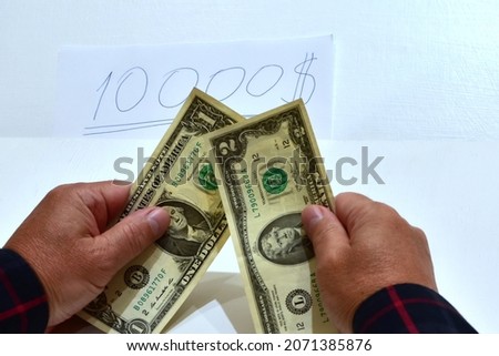 Elderly man hands hold 1 and 2 dollars banknotes against paper with written 10000 dollars on the table. Blurred foreground