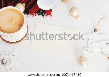 Nordic style Christmas composition. Flat lay blank greeting card mockup, cinnamon sticks, cup of coffee, bell, balls, knitted hat, wooden Christmas decorations on white table. Hygge, cozy home decor c