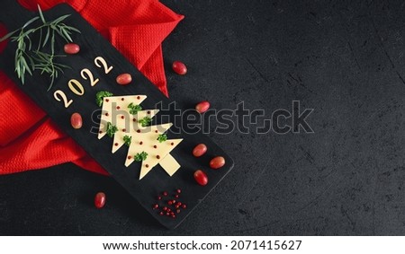 Creative Christmas tree made of cheese with herbs, spices, wooden numbers 2022 and grapes on a cutting board with a red kitchen napkin on the left on a black background with a place for text 